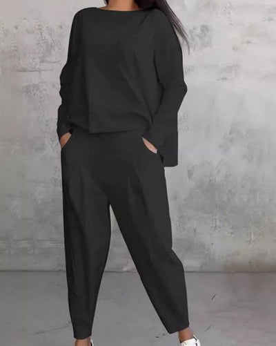 Femme en ensemble noir élégant, pose décontractée, mains dans les poches, fond gris minimaliste. Mode féminine, tenue chic, style moderne.