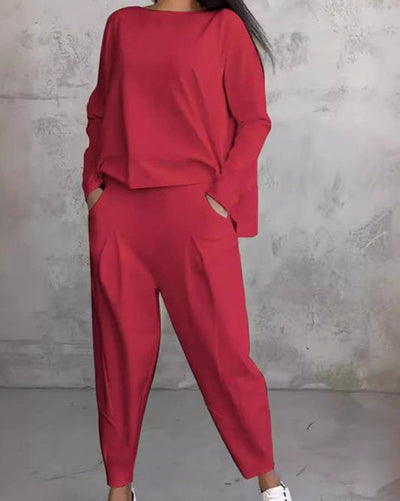 Femme portant un ensemble rouge élégant, composé d'un haut ample et d'un pantalon taille haute, posant contre un mur gris texturé.