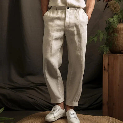 Pantalon en lin beige pour homme, style décontracté, avec ceinture, associé à des baskets blanches, ambiance naturelle avec plantes en arrière-plan.