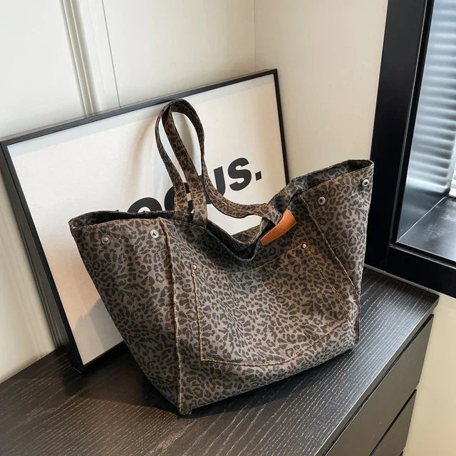 Sac à main en toile imprimé léopard, posé sur une table noire, avec un cadre minimaliste en arrière-plan. Mode, accessoire tendance, design chic.
