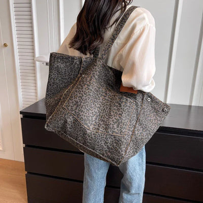 Femme portant un grand sac cabas à motif léopard, chemise blanche et jean, debout devant une commode noire dans une pièce moderne.