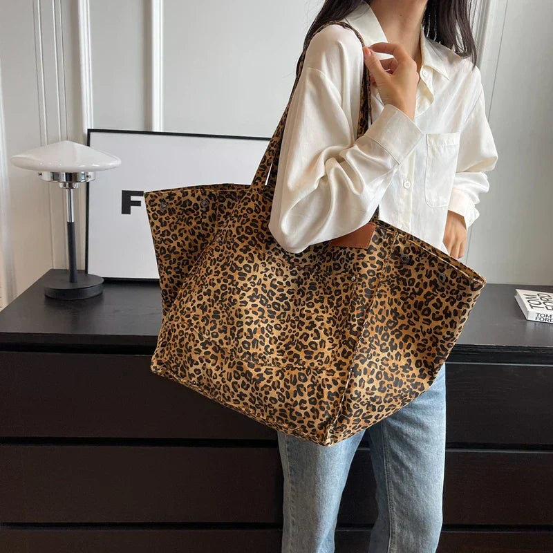 Femme portant un grand sac à main imprimé léopard, chemise blanche et jean, devant une commode noire. Mode tendance, accessoire animalier.
