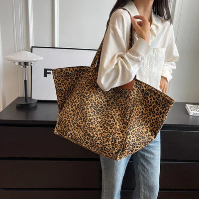 Femme portant un grand sac à main imprimé léopard, chemise blanche et jean, devant une commode noire. Mode tendance, accessoire animalier.