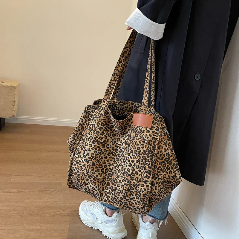 Sac à main imprimé léopard, porté par une personne en manteau noir et jeans, sur fond de sol en bois. Mode tendance, accessoire chic.