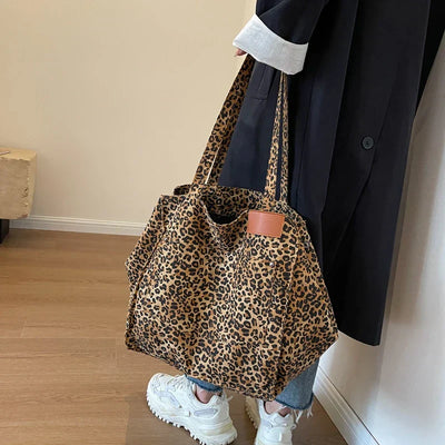 Sac à main imprimé léopard, porté par une personne en manteau noir et jeans, sur fond de sol en bois. Mode tendance, accessoire chic.