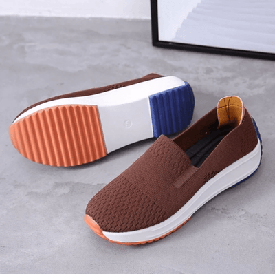 Chaussures marron en toile avec semelle blanche et détails orange et bleu, posées sur un sol gris. Design moderne et confortable pour un style décontracté.