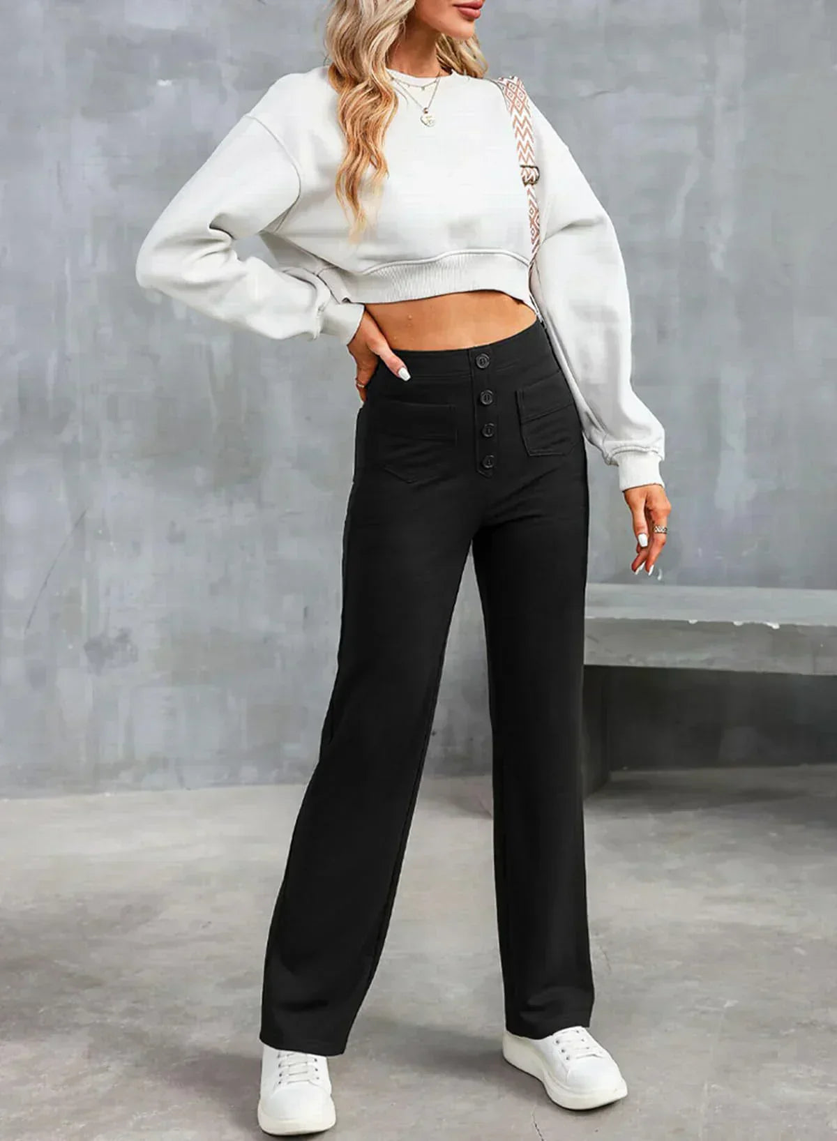 Femme portant un pull blanc court et un pantalon noir taille haute avec boutons, posant contre un mur gris. Mode décontractée, tendance automne.