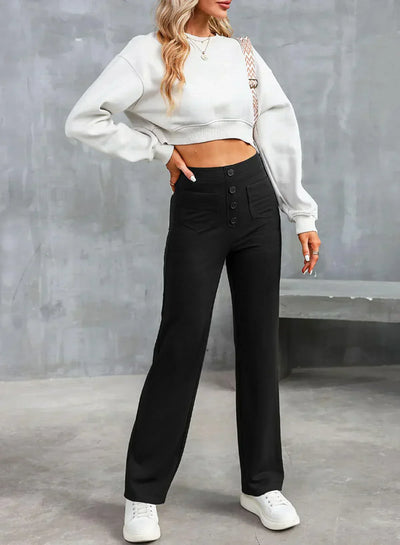 Femme portant un pull blanc court et un pantalon noir taille haute avec boutons, posant contre un mur gris. Mode décontractée, tendance automne.